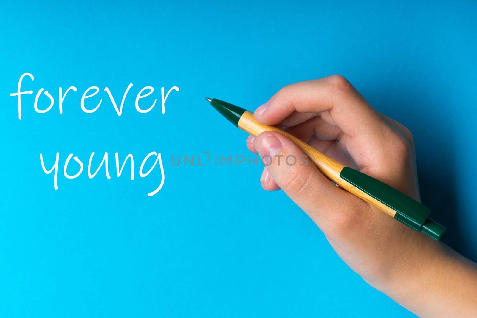 Children hand with pen write on a blue white background. Writing hand. Word Forever young