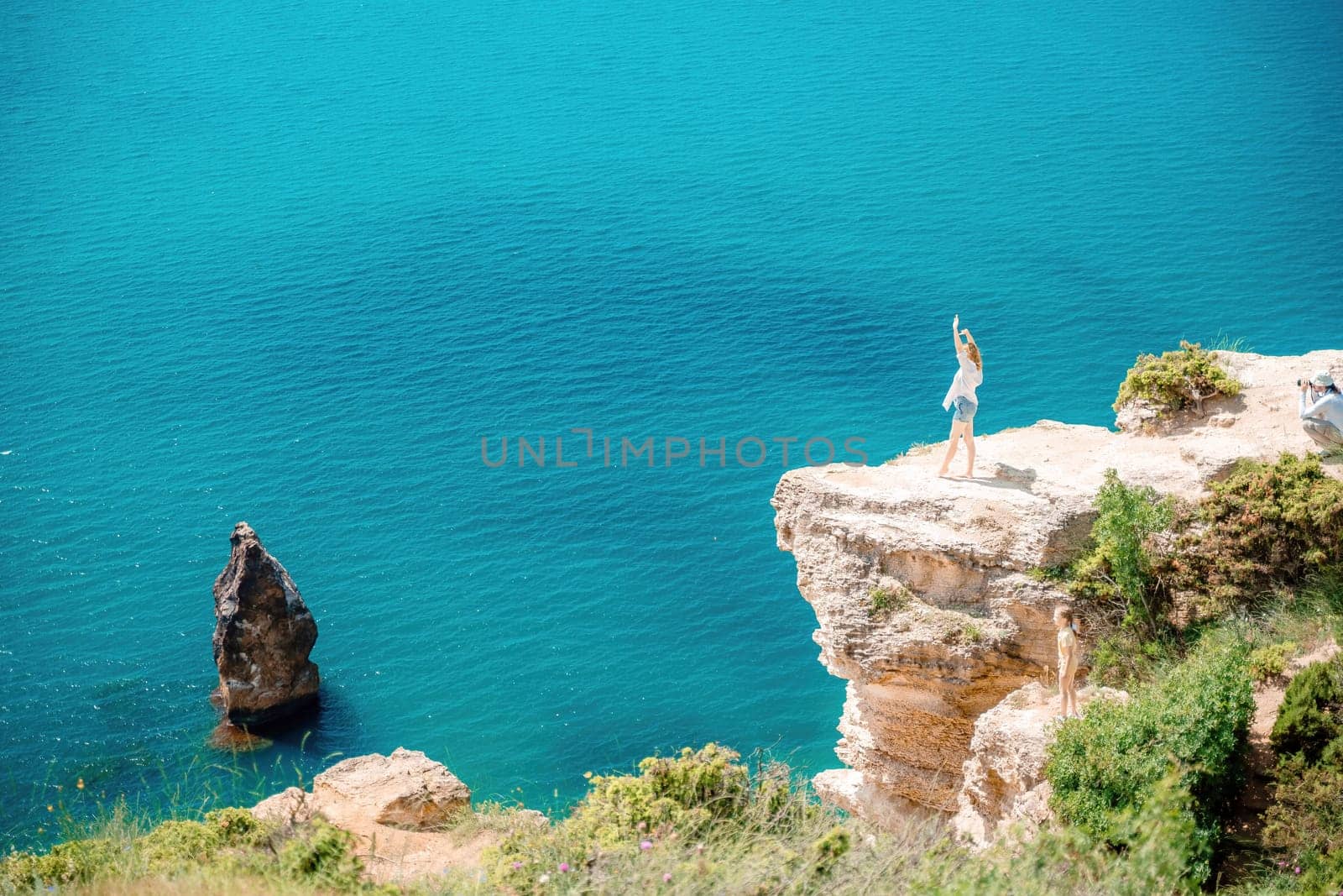 A woman stands on a cliff overlooking the ocean. The water is blue and the sky is clear