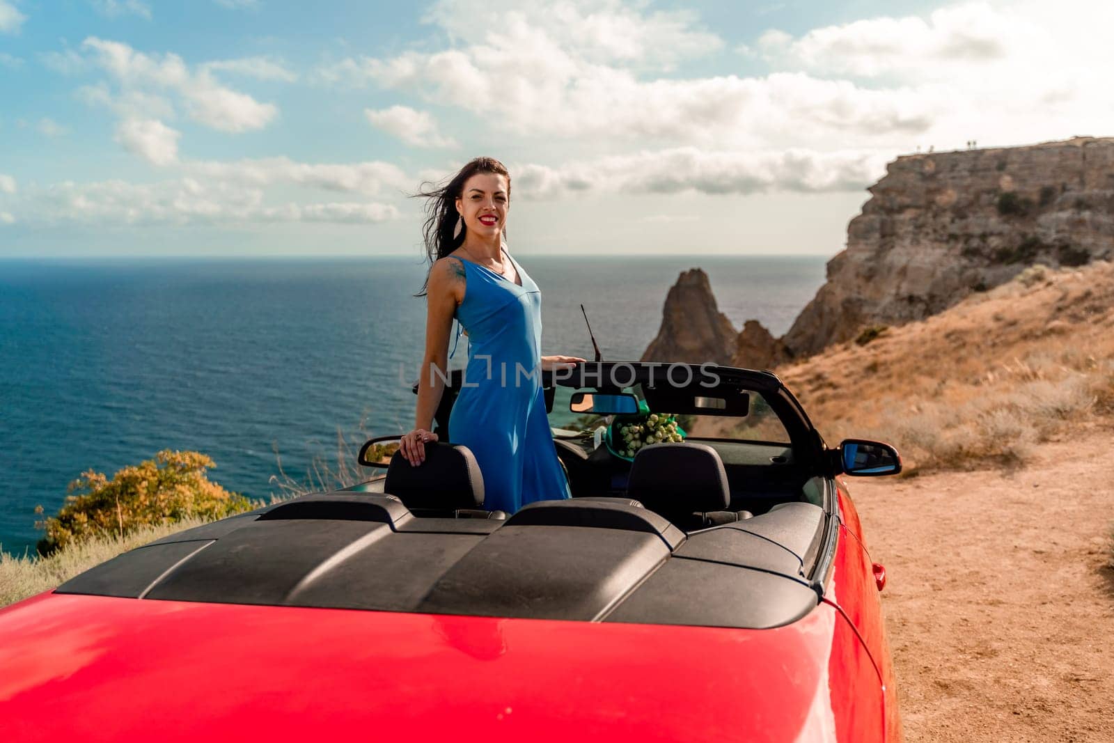 A woman is standing in front of a red convertible car. She is wearing a blue dress and smiling. The scene is set on a beach, with the ocean in the background. Scene is cheerful and relaxed