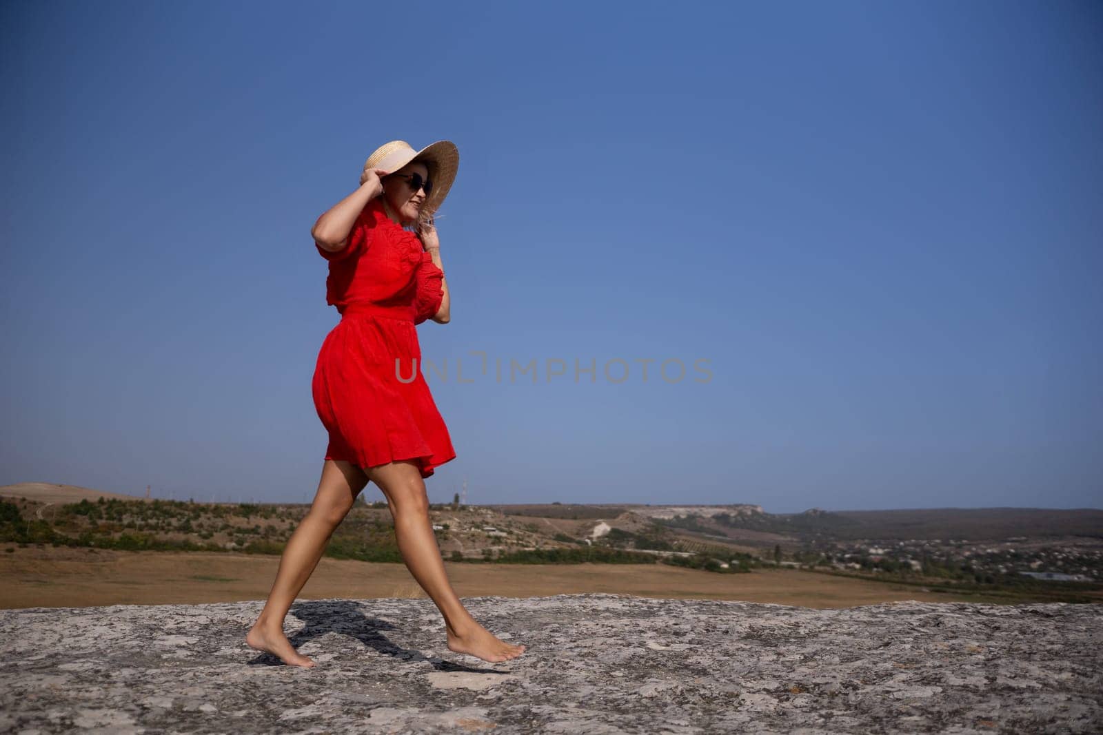 A woman in a red dress is walking barefoot on a rocky hillside. The sky is clear and blue, and the woman is wearing a straw hat. The scene is peaceful and serene