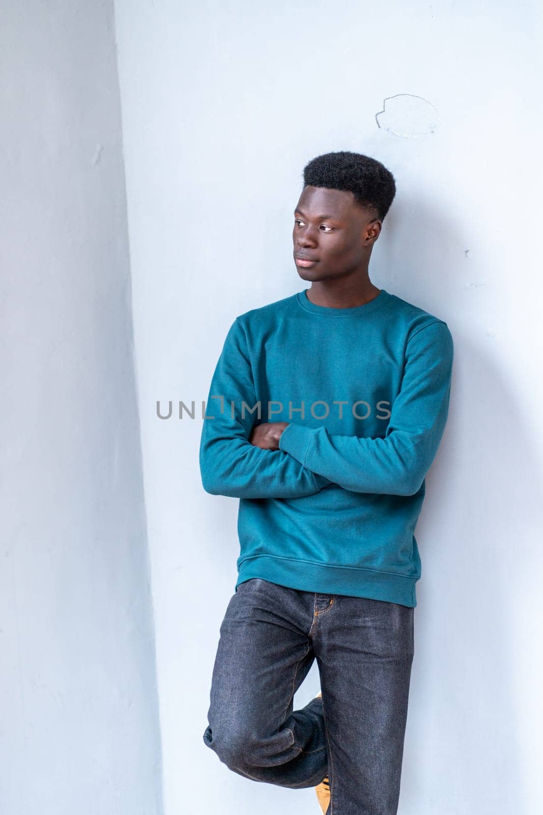 A young man in a blue sweatshirt and jeans stands against a white wall. He is looking off into the distance with his arms crossed. Concept of introspection and contemplation