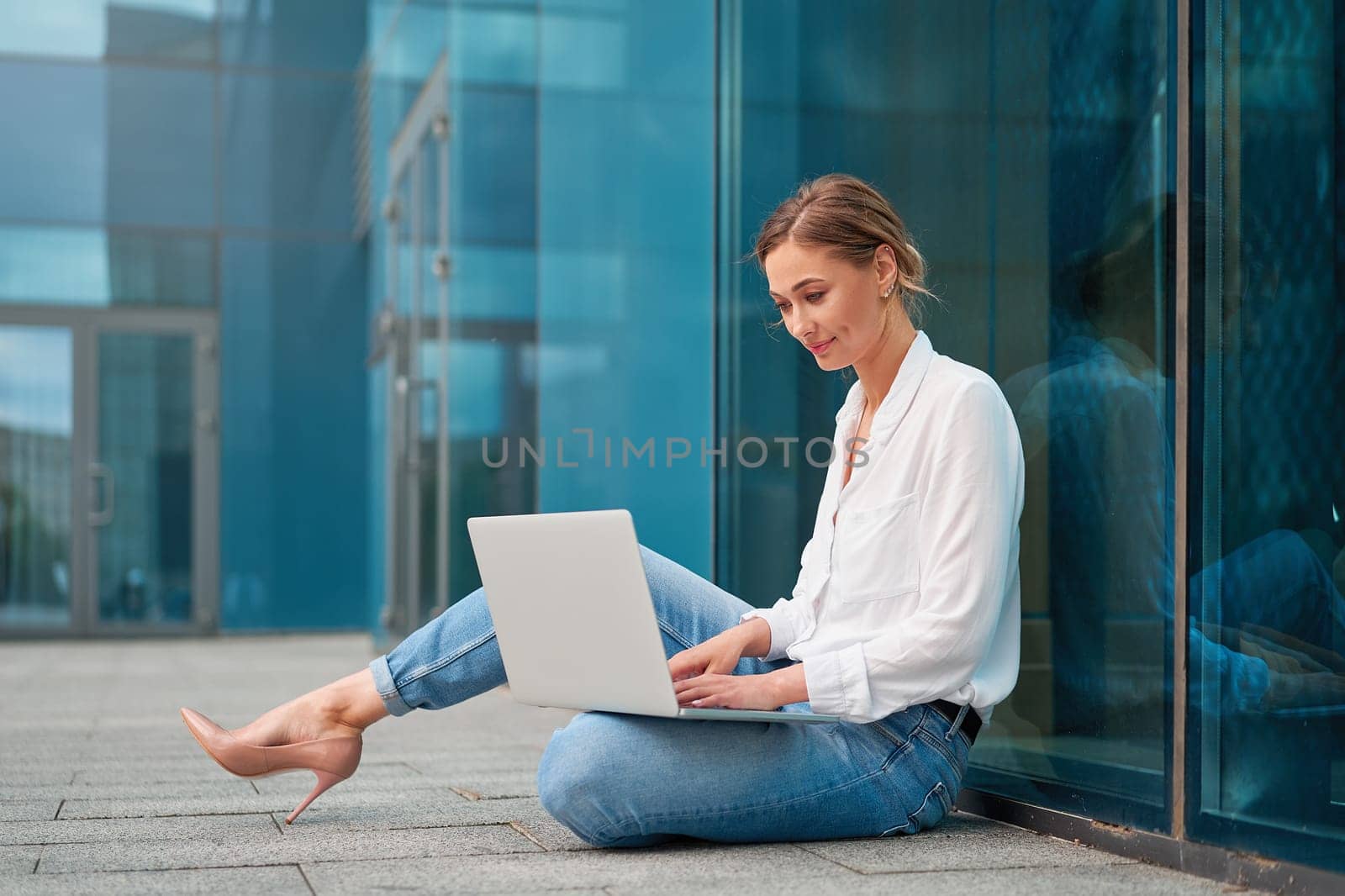 Full body side view of young businesswoman using laptop sitting on ground outside modern office. Confident female entrepreneur in smart casual working on netbook against glass building.