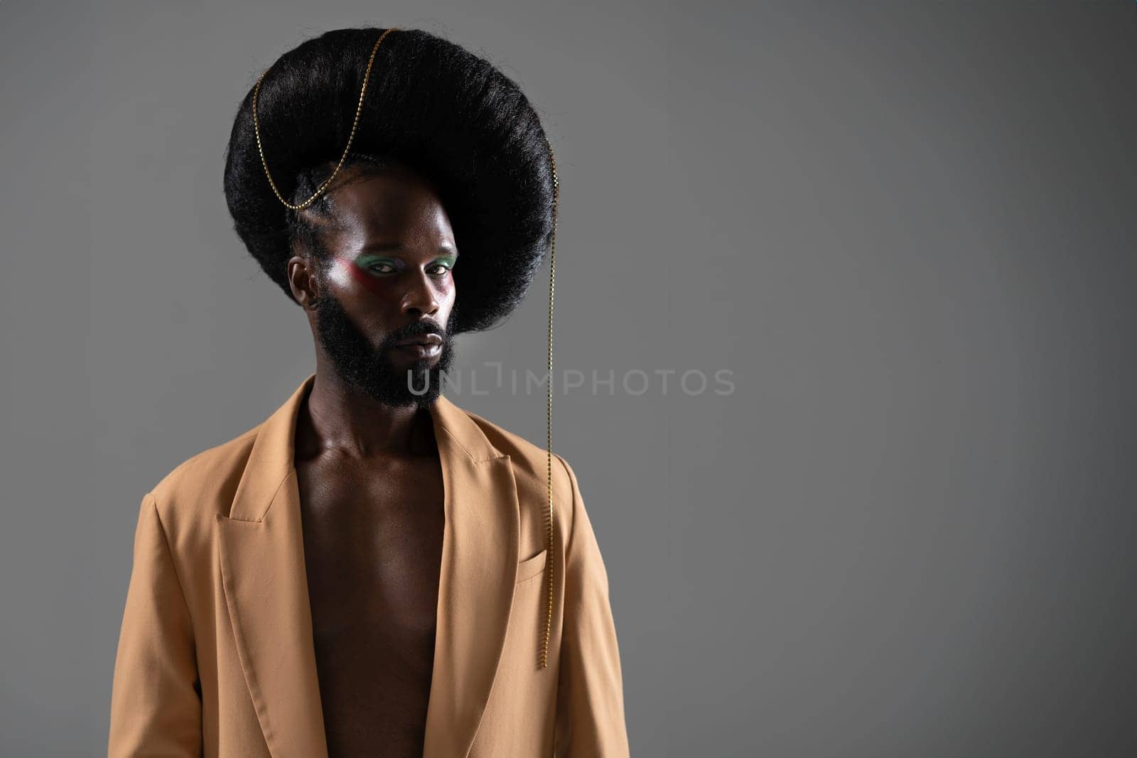 Portrait of fashionable African-American bearded gay man with light makeup and unique retro wig hairstyle, isolated on gray background. He is dressed in beige jacket on shirtless body.
