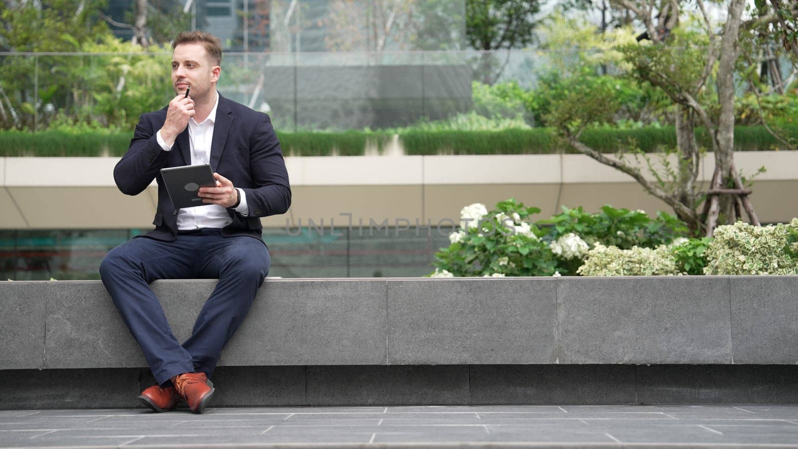 Caucasian smart businessman sitting at park while writing on tablet by using tablet pencil and planning marketing strategy. Manager looking at data analysis while thinking about financial plan. Urbane