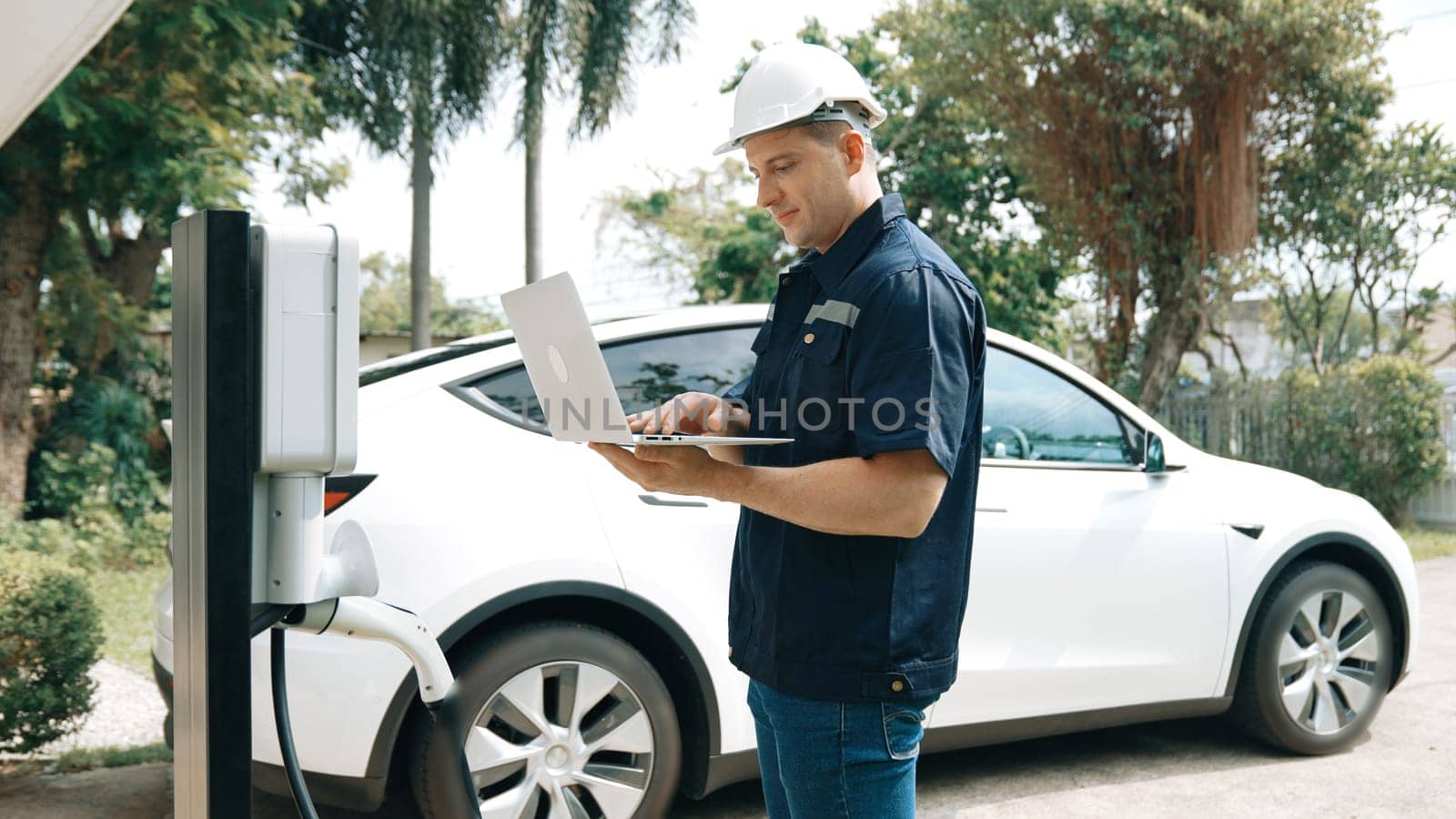 Qualified technician working on home EV charging station installation, making troubleshooting and configuration setup on charging system with laptop for EV at home. Synchronos