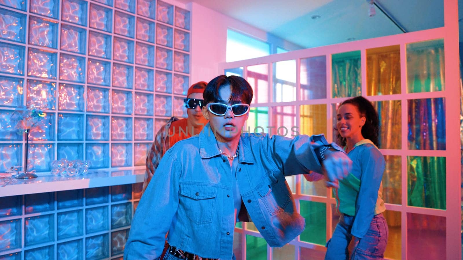 Asian man moving his arm while dancing with diverse team in neon light at night club. Happy dancer with stylish cloth moving along with multicultural hipster in lively mood at modern club. Regalement.