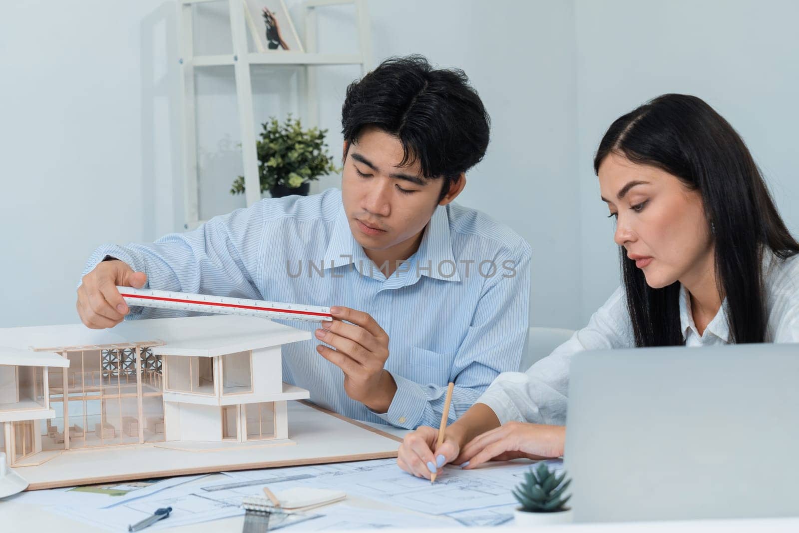 Professional male asian architect using ruler to measure house model length while young beautiful caucasian colleague using laptop to analyzed data on meeting table with house model. Immaculate.