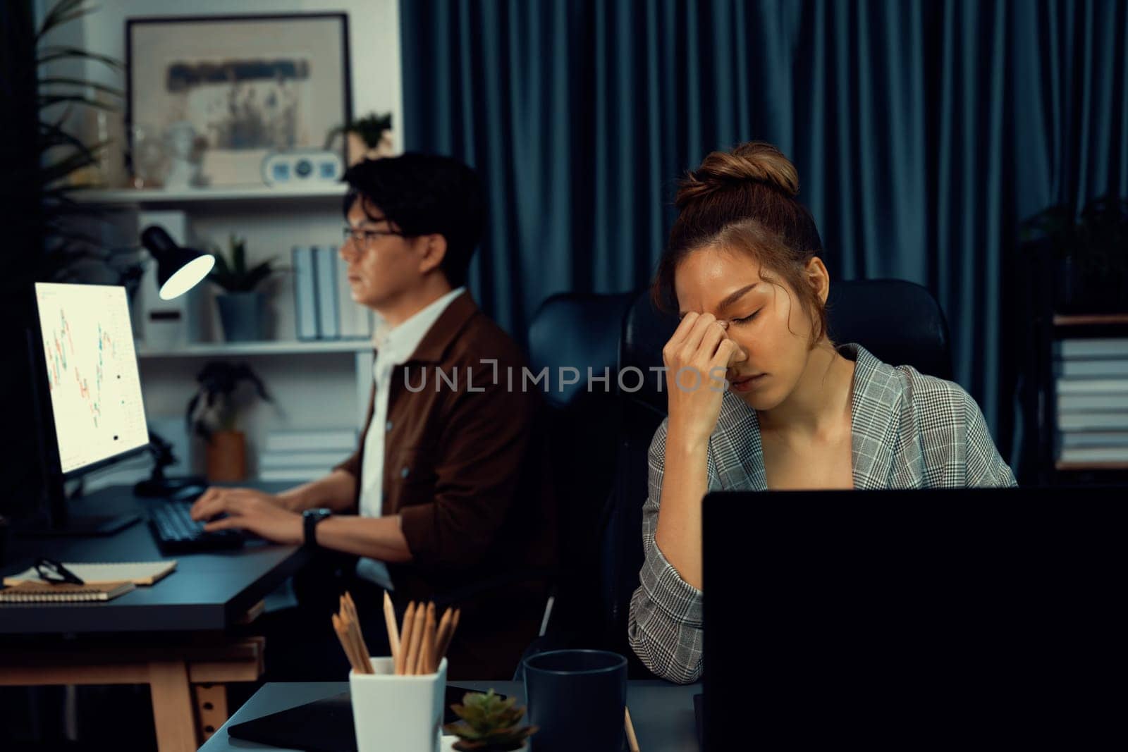 Businesswoman working on desk with stretching arm up and down manner with body health ache of strain overwork while coworker trading stock market on website on pc at late over night time. Infobahn.