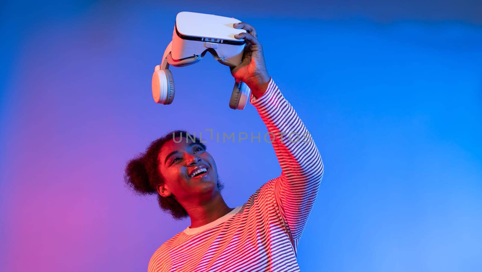 Smiling enthusiastic young African woman looking at VR headset surprising through metaverse world at vibrant neon pink and blue studio lighting background technology learning cyberspace. Contrivance.