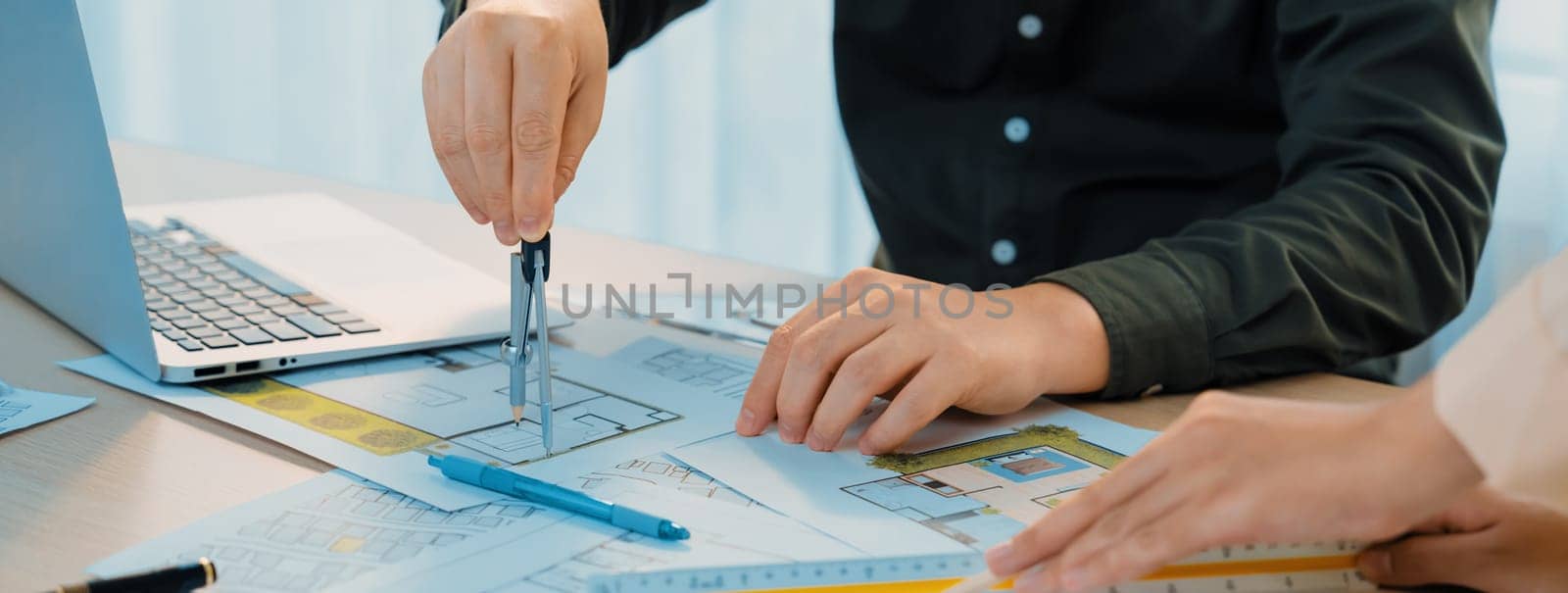 A cropped image of professional engineer using divider to measure blueprint at meeting table with blueprint, laptop and architectural equipment scatter around. Closeup. Delineation.