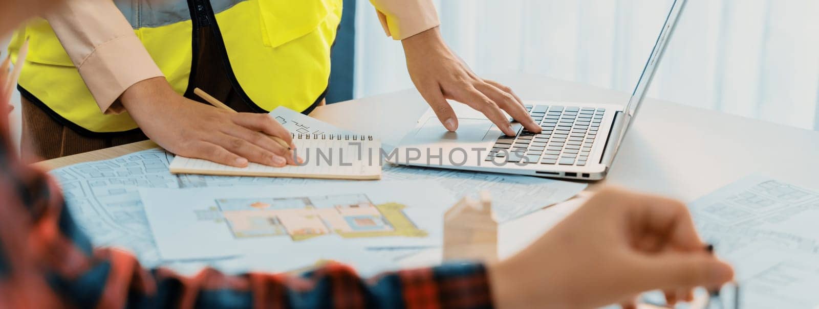 Cropped image of professional engineer team working on blueprint while his coworker working on laptop at meeting table with blueprint and wooden block scattered around. Closeup. Delineation.