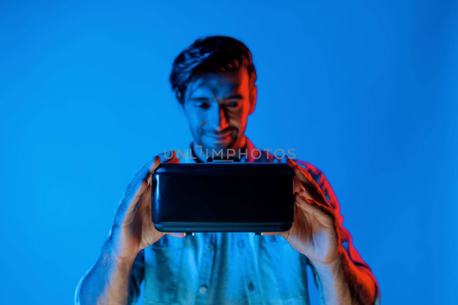 Smiling caucasian man holding and show VR headset with blurring background while standing with neon light. Smart teenager looking at digital goggles to enter virtual reality or metaverse. Deviation.