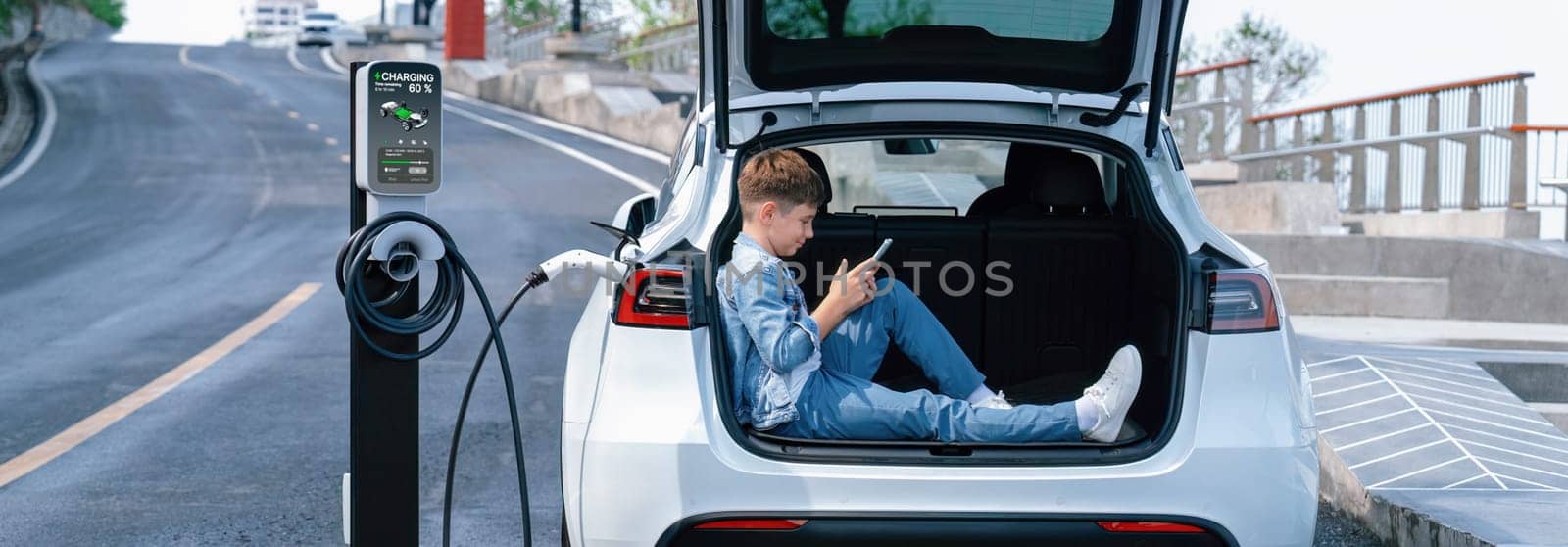 Little boy sitting on car trunk, using smartphone while recharging eco car from EV charging station. EV car road trip travel as alternative vehicle using sustainable energy concept.Panorama Perpetual