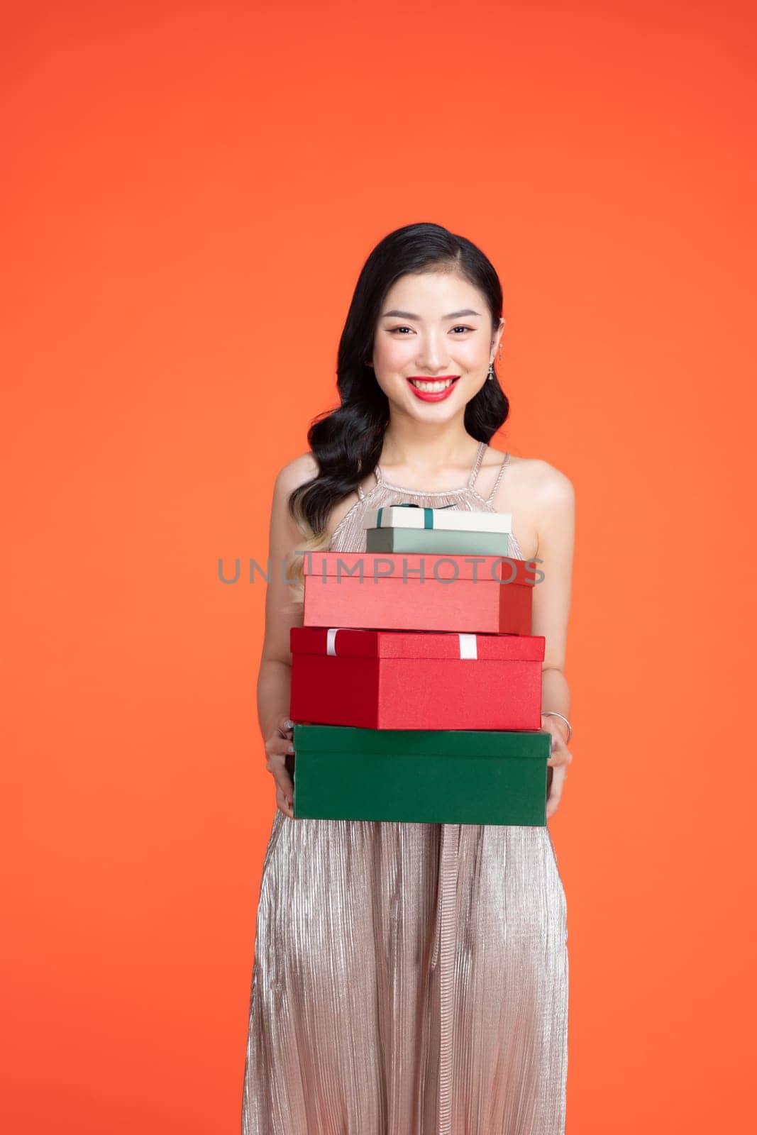 Charming female wearing shiny dress standing with pile of celebrating presents on red background