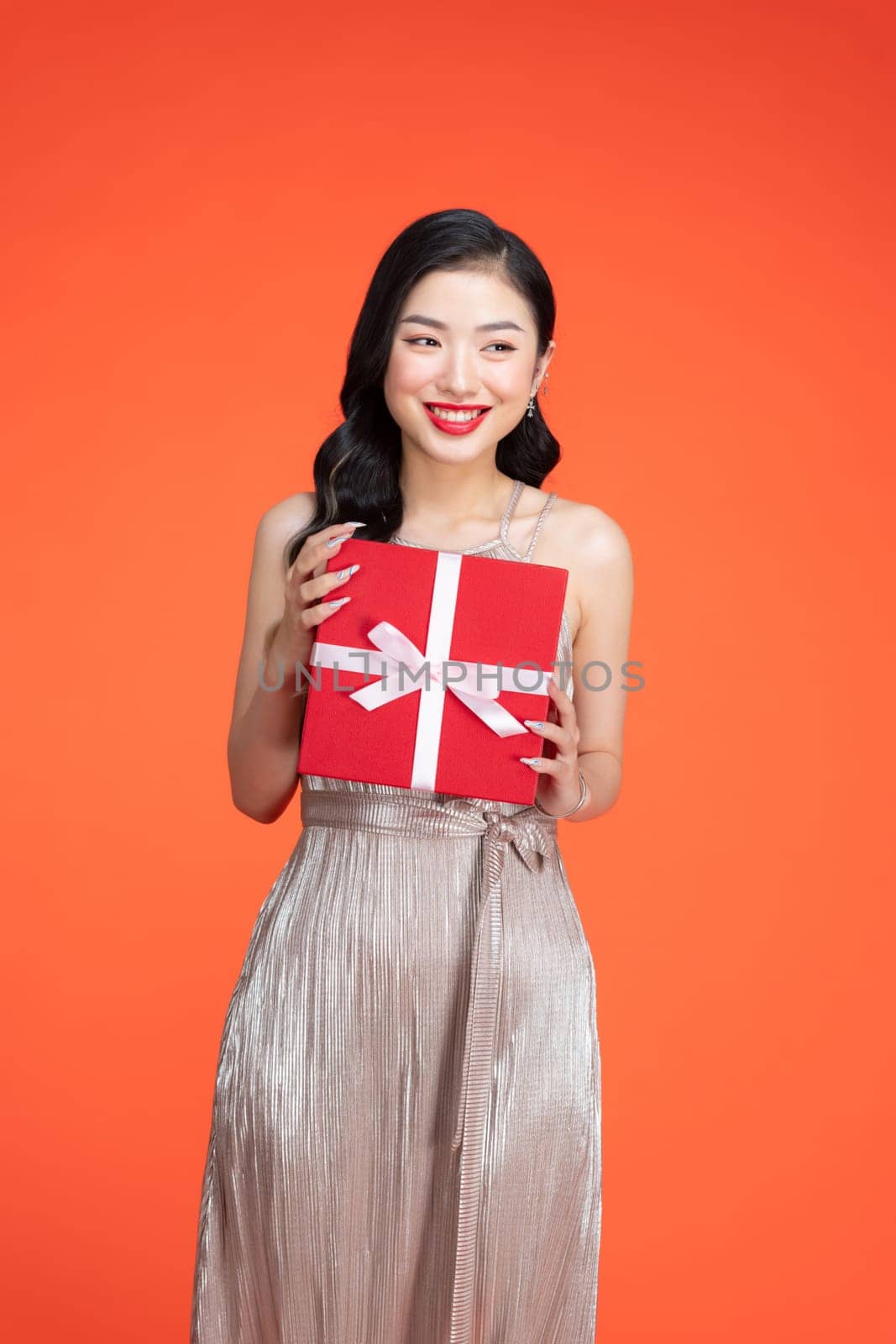 Happy dreamy lady in elegant golden dress holding wrapped gift box, and smiling, posing on red background