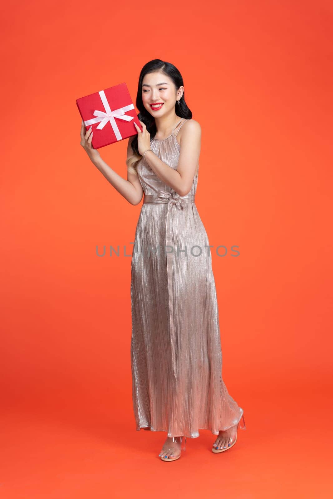 Portrait of happy girl shaking gift box wondering what inside as celebrating presents on red background by makidotvn