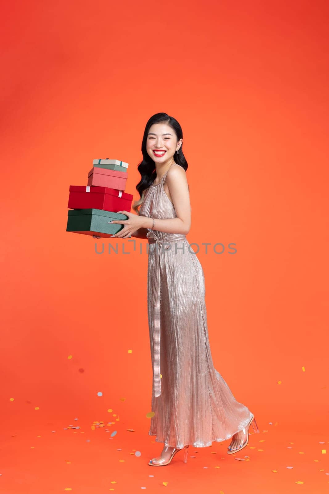 Cheerful young woman in shiny dress holding heap of presents, looking aside on red background