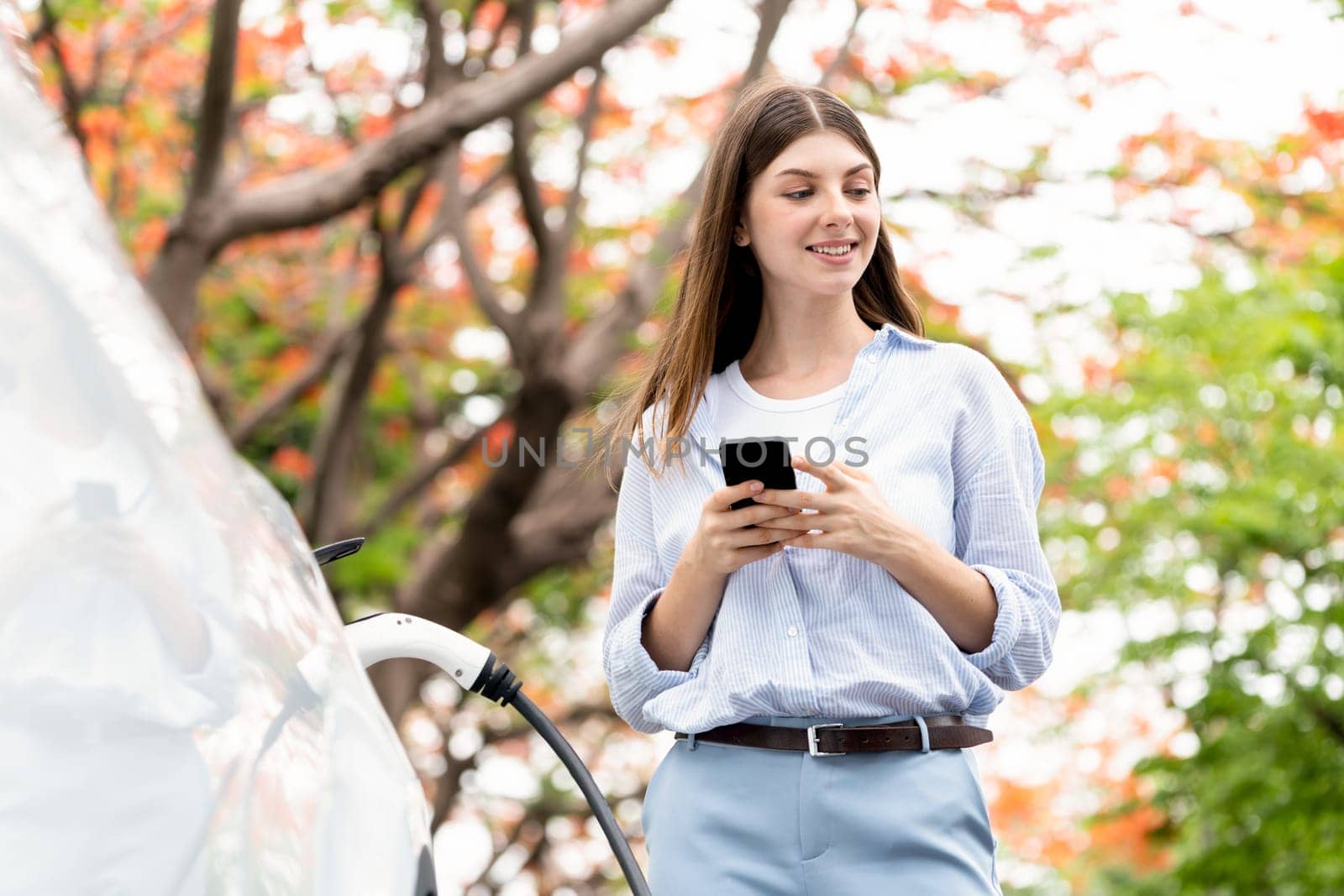 Young woman using smartphone online banking application to pay for electric car battery charging from EV charging station during autumn vacation holiday trip at national park or autumnal forest. Exalt