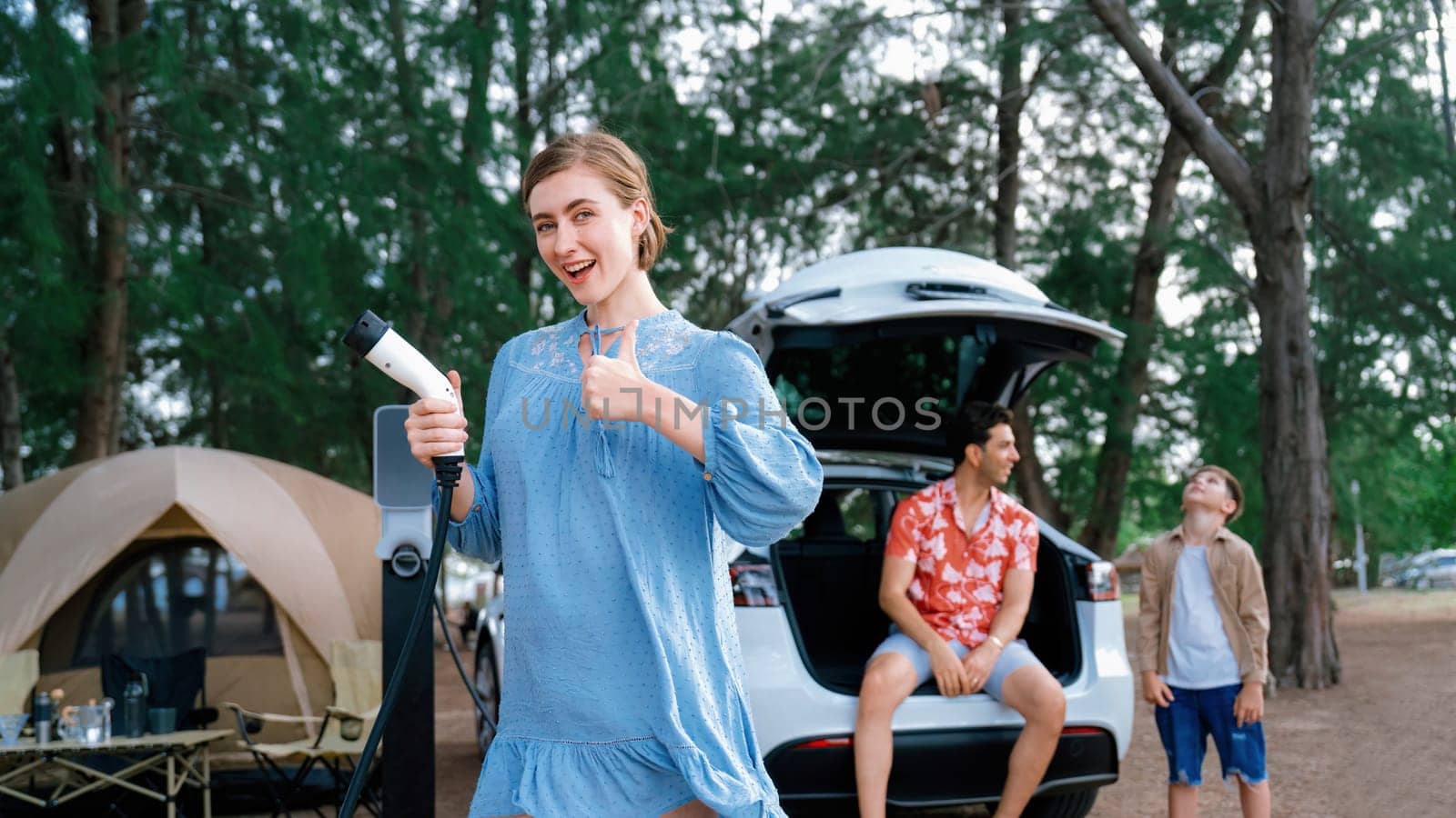 Outdoor adventure and family vacation camping at sea travel by eco friendly car. Cheerful woman or mother holding, pointing EV charger point with playful and happiness posture in campsite. Perpetual