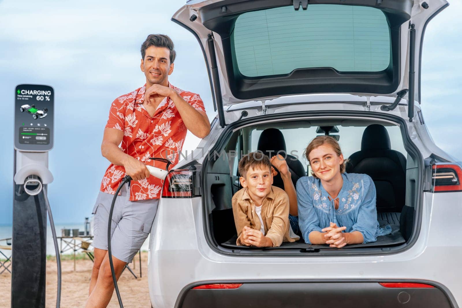 Family vacation trip traveling by the beach with electric car, lovely family sit on the trunk, charging EV car battery with green and sustainable energy. Family travel and eco-friendly car. Perpetual