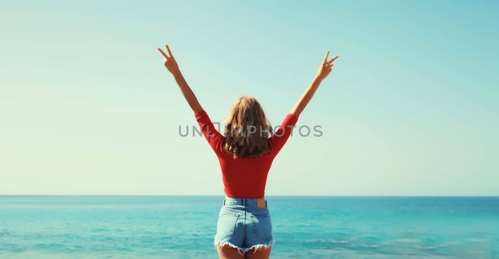 Summer vacation, rear view of happy inspired young woman raising her hands up on the beach on sunny sea coast and blue sky background