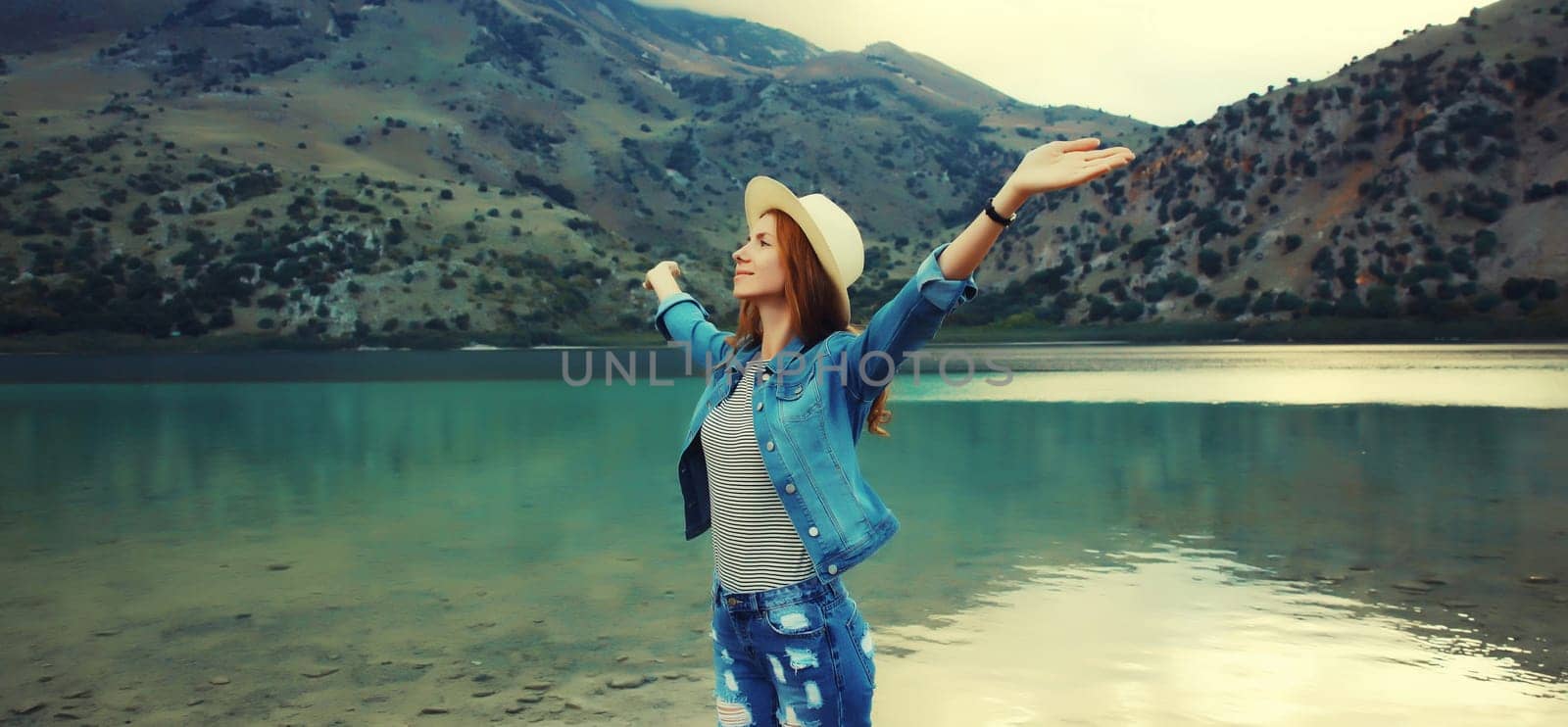 Summer vacation and tourism, rear view of happy woman tourist in straw hat raising her hands up on lake and mountains background. Greece, island Crete.