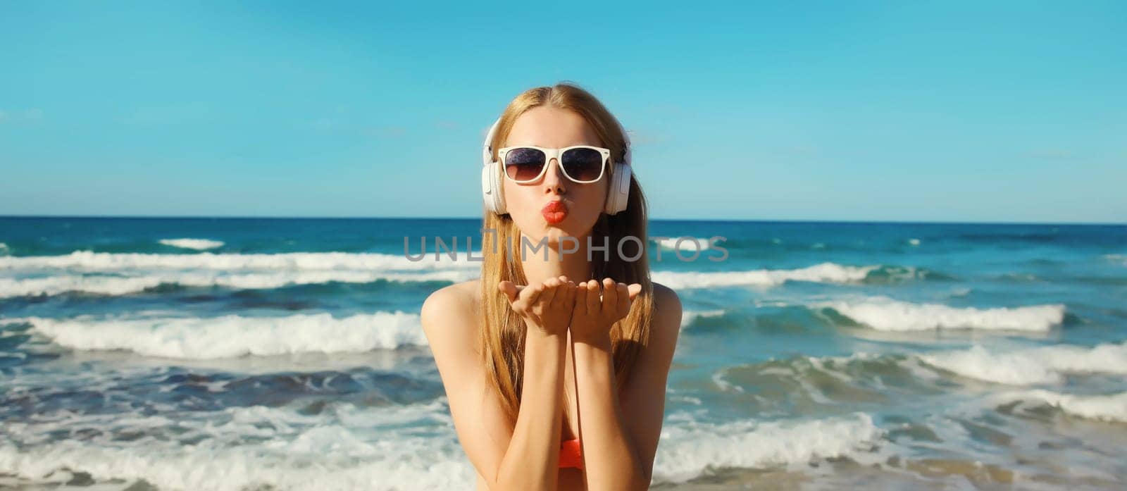 Summer vacation of happy young woman relaxing listening to music with headphones, modern girl blowing a kiss having fun on the beach at sea coast with waves on a sunny day