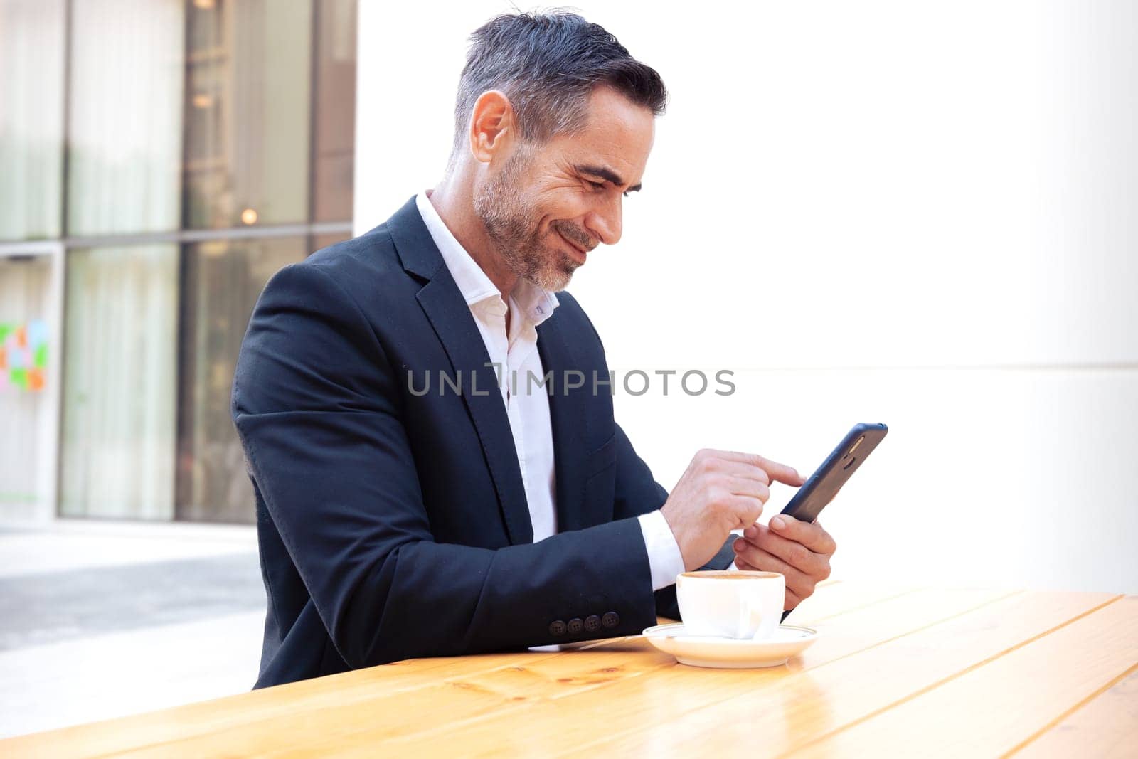 Satisfied with the results, the businessman sitting outside the office building, a mature boss holds a phone and drinks coffee, writes messages and reads news online, using an app.