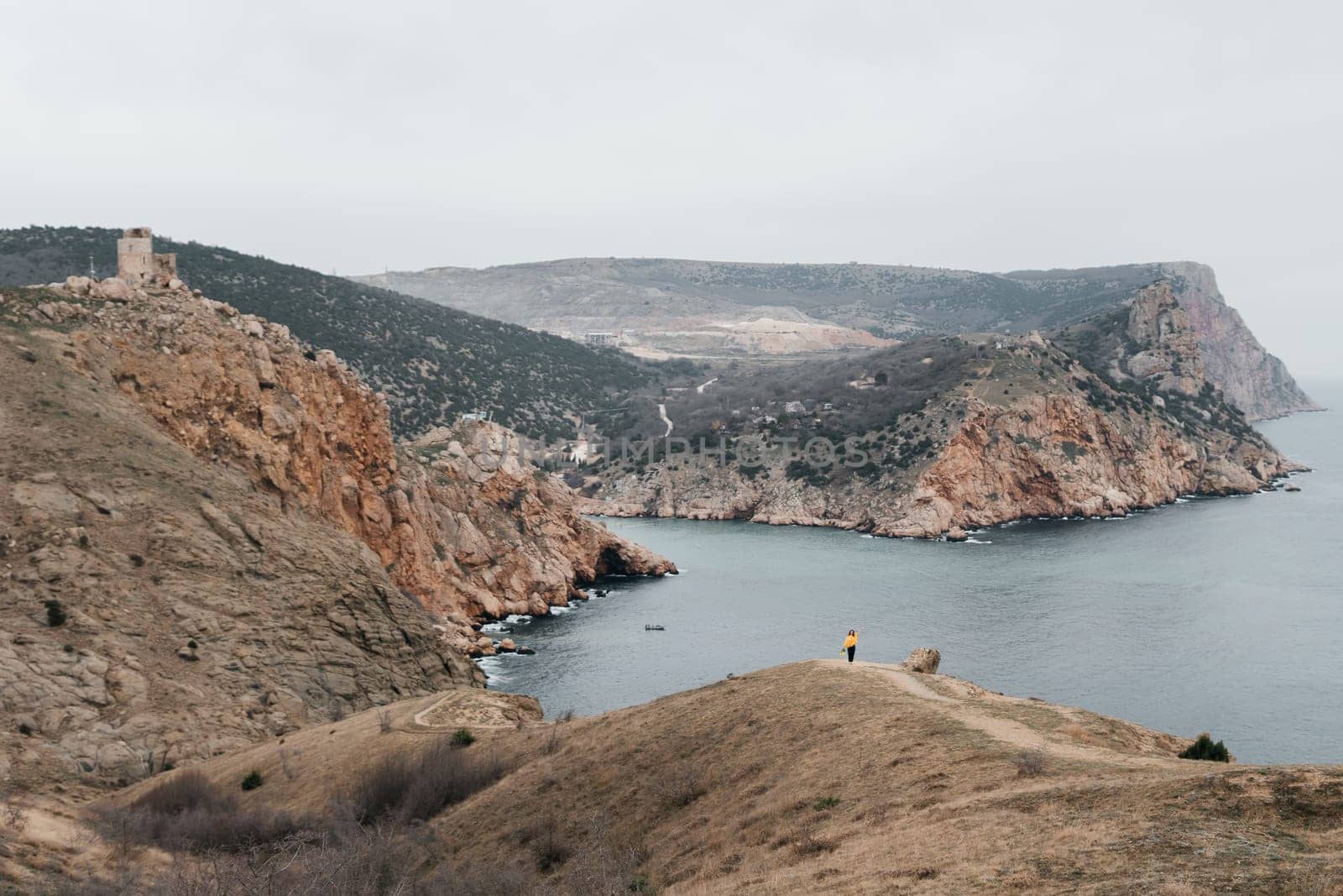 Woman stands on cliff overlooking ocean, cloudy day provides soft lighting. She's wearing a yellow sweater wearing dark pants. Landscape features rugged terrain, calm waters