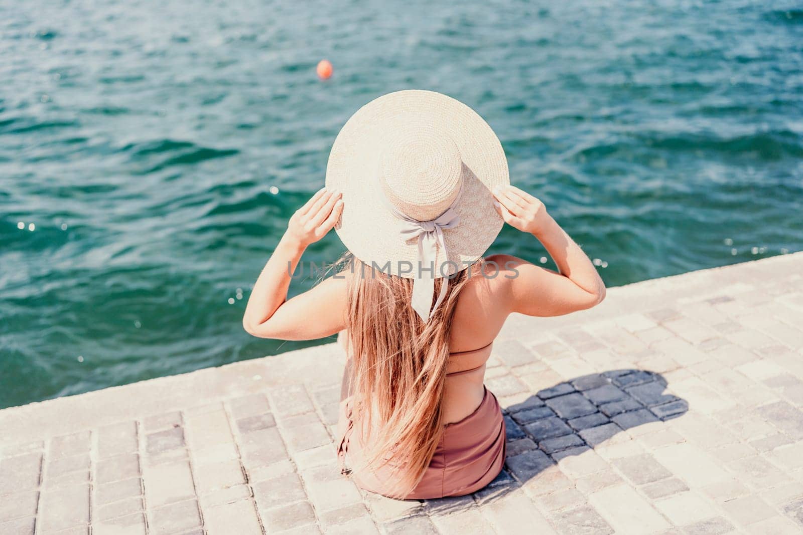 A woman in a swimsuit sits with her back holding a hat, looks at the ocean, sunny day, relaxes