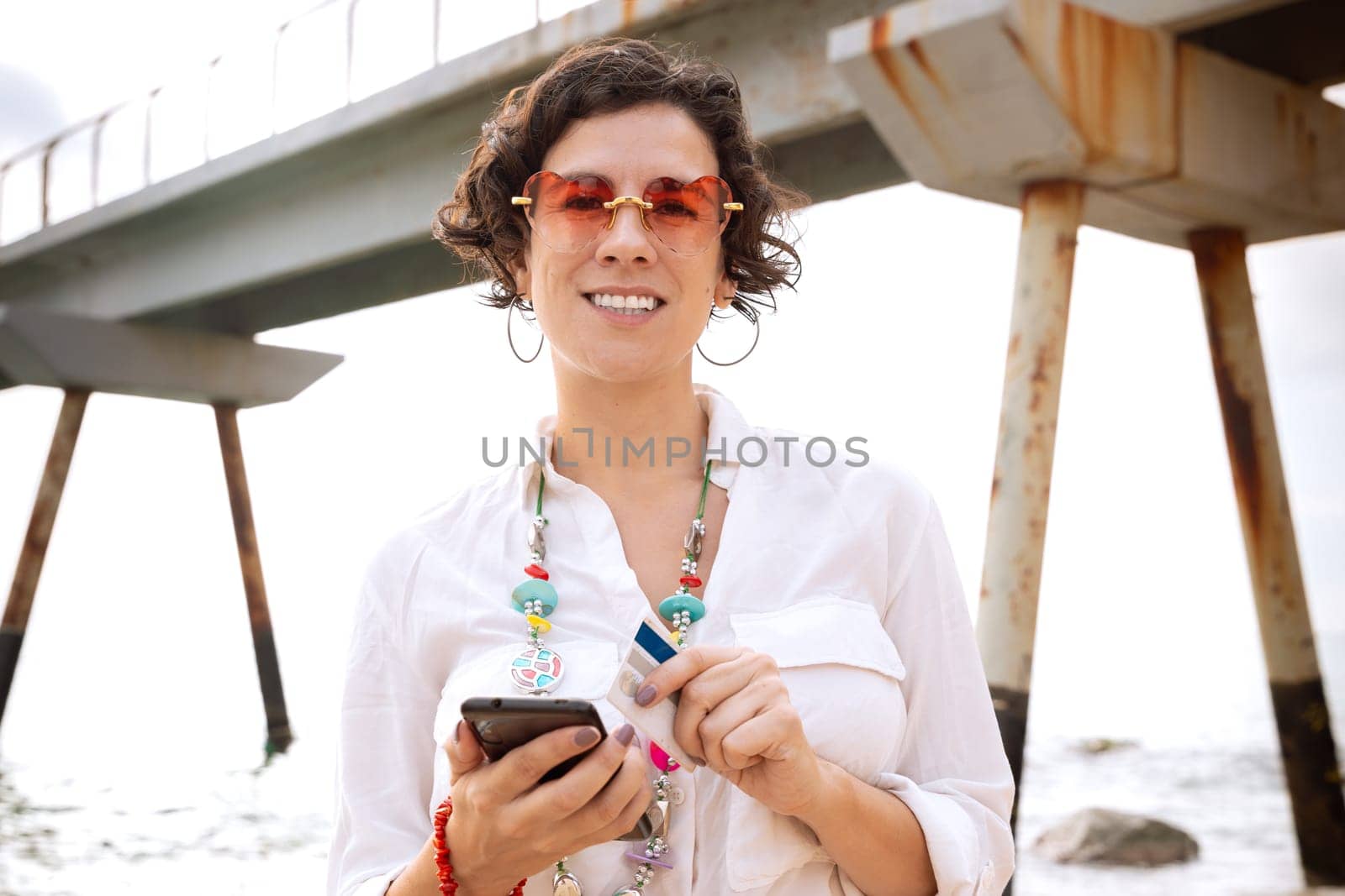 caucasian woman wearing sunglasses on vacation by the sea laughing using a social media app with her mobile phone,