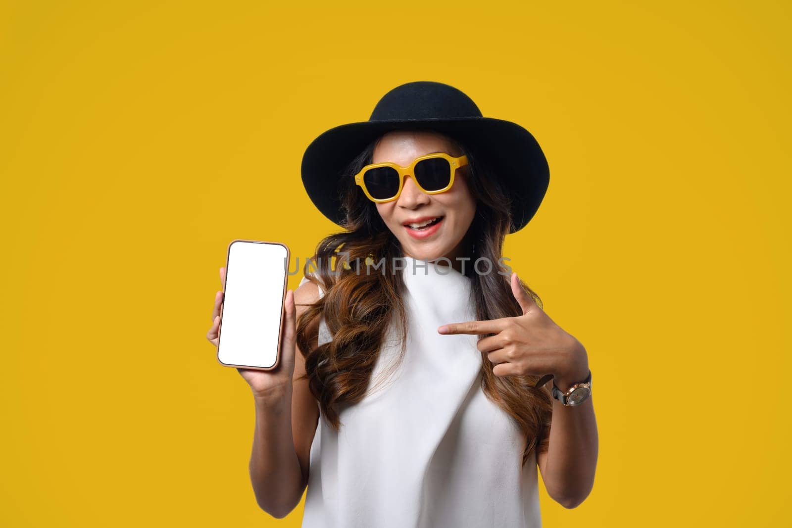 Smiling female traveler standing isolated over yellow background and showing mobile phone for presenting.