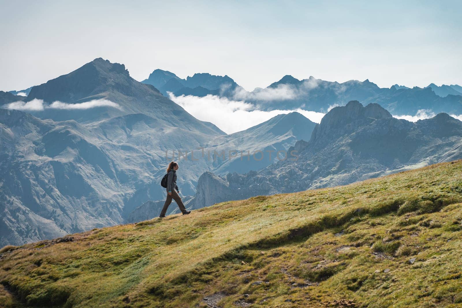 A person is walking on top of a grassy hill in the mountains, enjoying the natural landscape, surrounded by the beauty of the sky, clouds, and mountain range