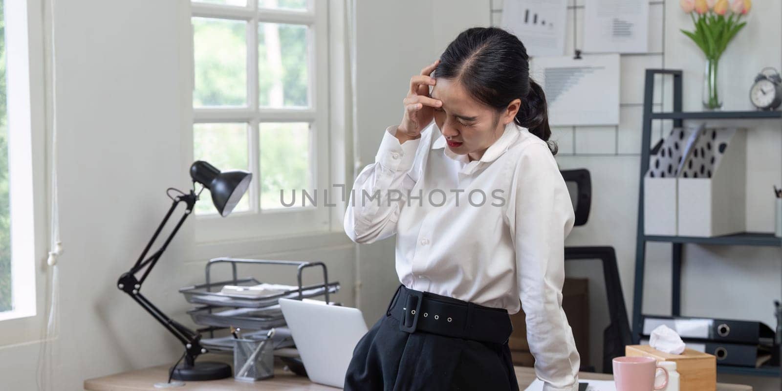 Businesswoman in office suffering from headache, surrounded by documents, laptop, and office supplies, depicting workplace stress.