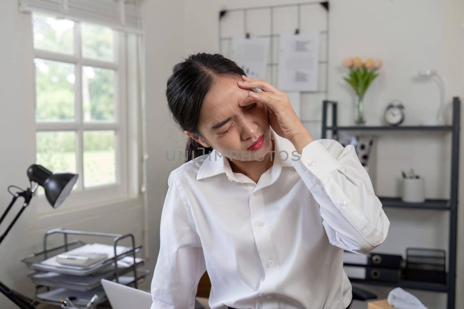 A businesswoman in a white shirt experiencing stress and headache in an office setting, surrounded by documents and a laptop.