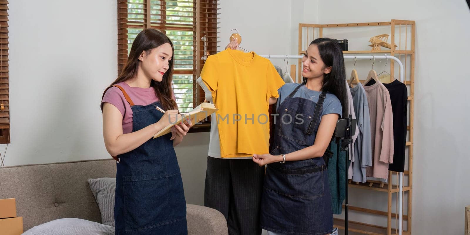 A lesbian couple conducting an online live selling session from home, displaying clothing items and managing their business.