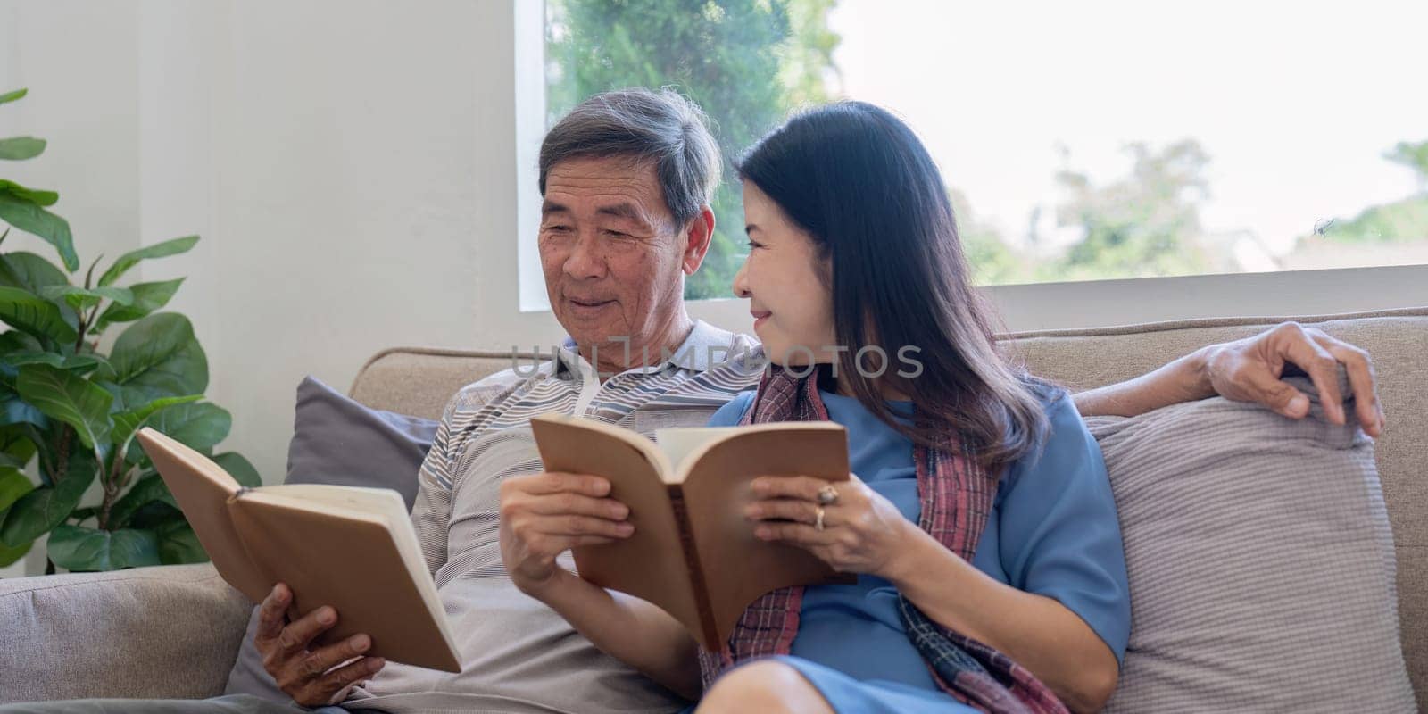 An elderly couple sits together on a comfortable sofa, enjoying reading books in a bright and cozy living room.