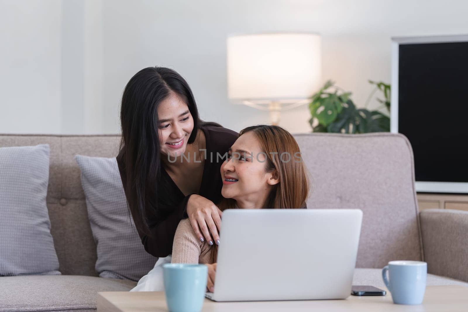 Happy LGBT Lesbian Couple Sitting Together on Couch with Laptop in Cozy Living Room by wichayada