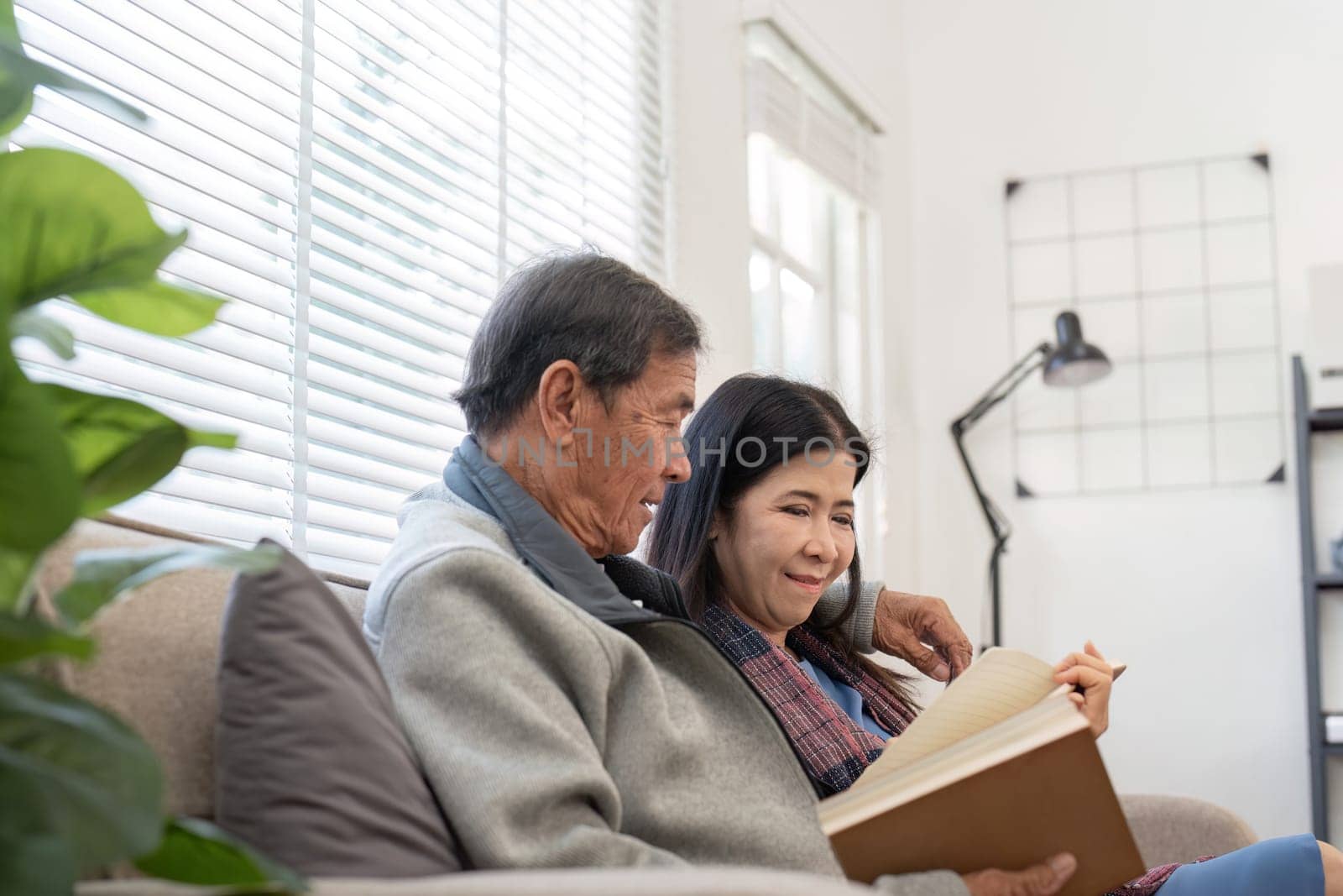 An elderly couple sits together on a sofa, sharing a book and enjoying each others company in a bright, modern living room.