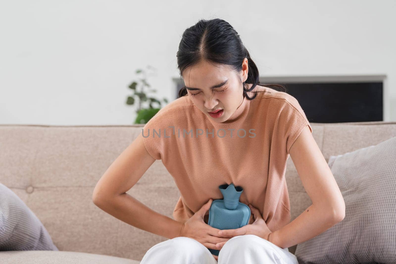 A woman sitting on a couch, holding a hot water bottle to her abdomen, experiencing menstrual pain and discomfort.