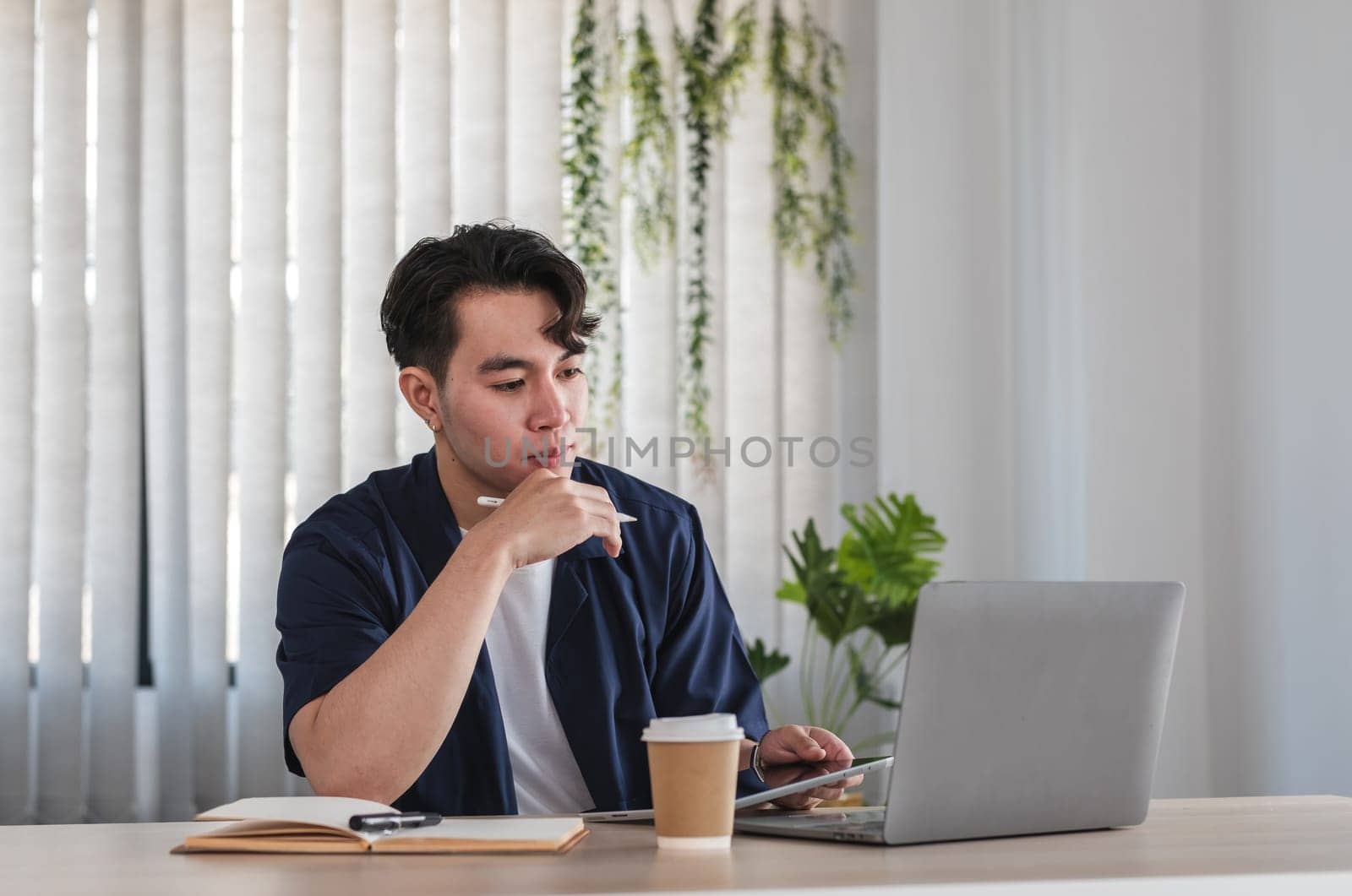 A young male businessman sits at a desk in a home office, working online on a laptop with a coffee cup nearby, appearing focused and thoughtful.