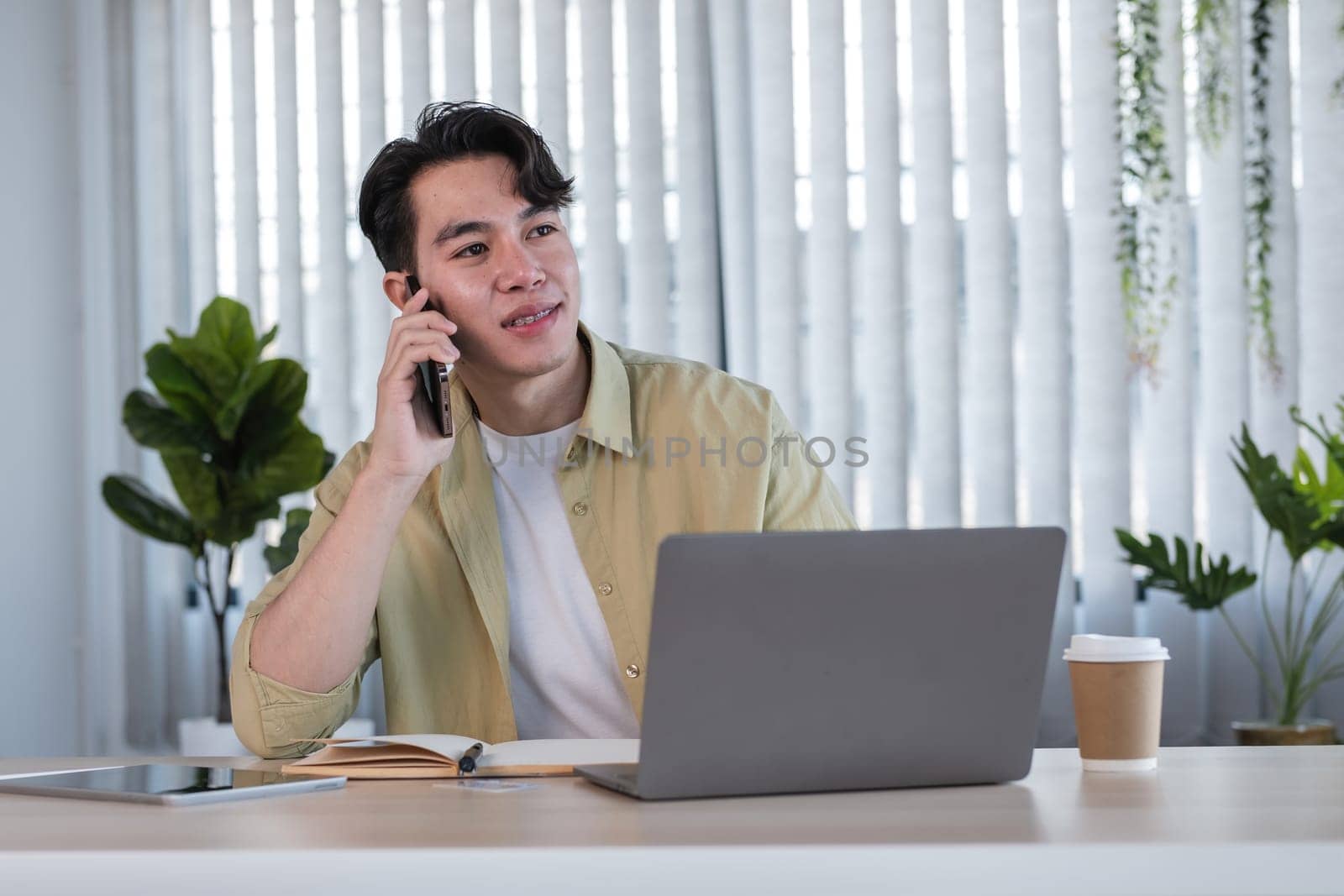 Young Male Businessman Working Online at Home Office, Using Laptop and Smartphone, Surrounded by Plants and Modern Decor by wichayada