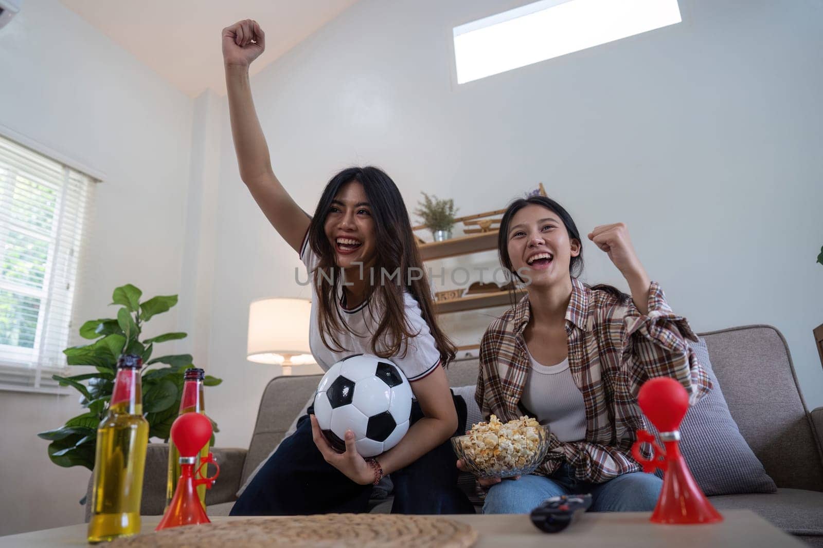 Happy lesbian couple celebrating a football victory at home, cheering with excitement and joy, holding a soccer ball and snacks.