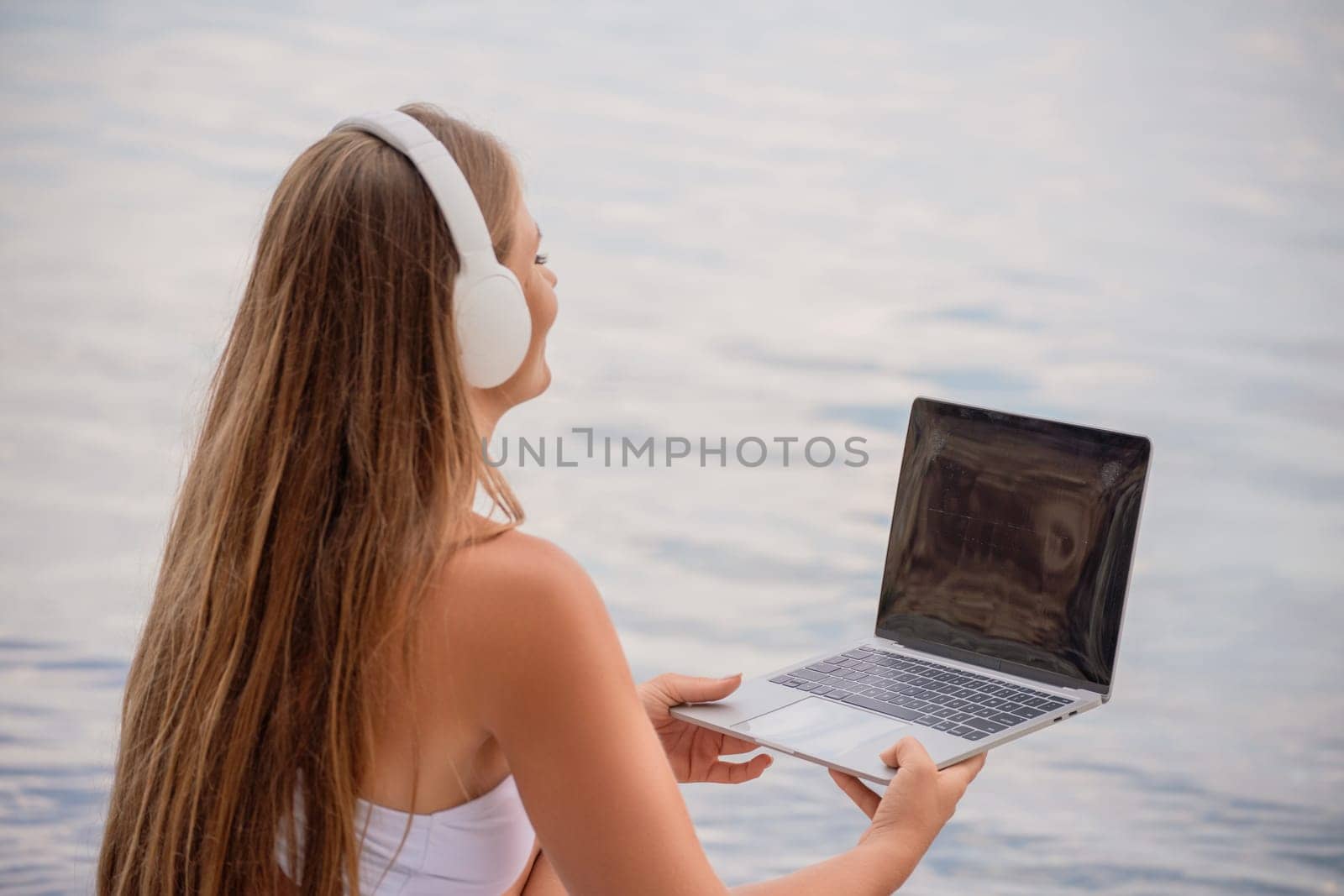 A woman is sitting by the water with a laptop in her lap. She is wearing headphones and she is listening to music. Concept of relaxation and leisure. by Matiunina