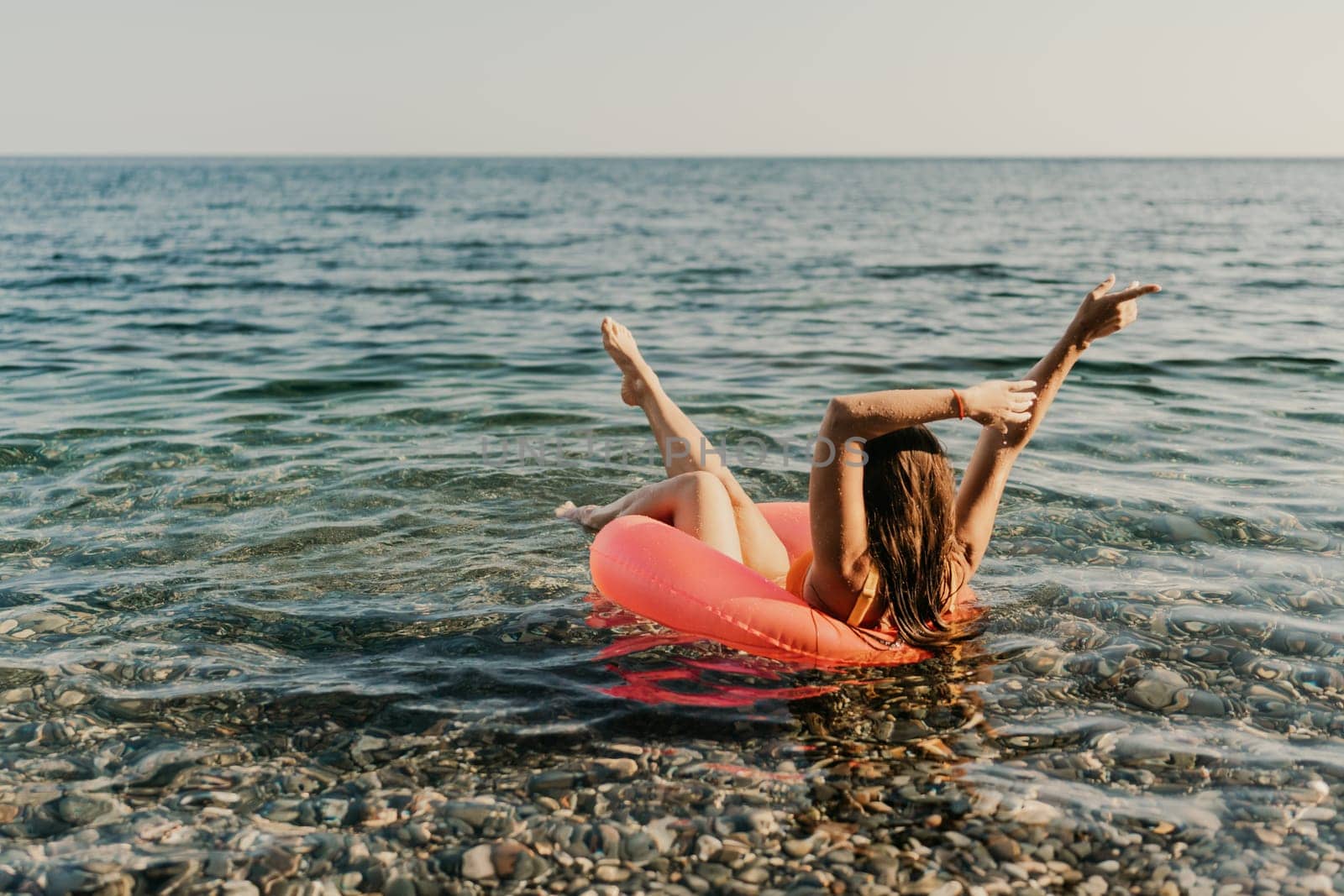 A woman is floating in a red inflatable raft on a rocky beach. The water is calm and the sky is clear. The woman is enjoying her time in the water, and the scene conveys a sense of relaxation