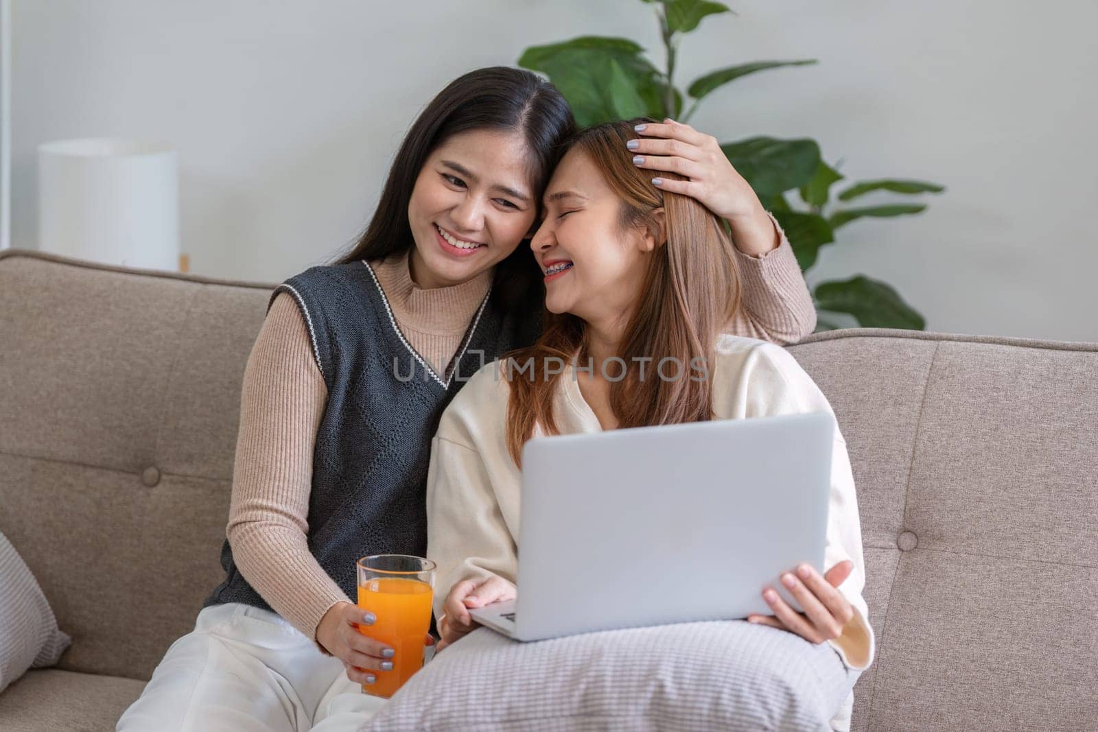 A joyful lesbian couple drinks fresh orange juice at home, sharing a moment of relaxation on the couch with a laptop.