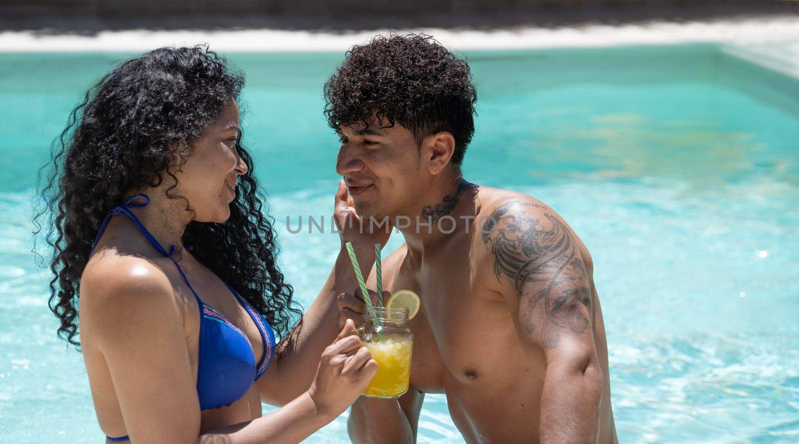 Young dominican couple are flirting on their first date in a turquoise water pool drinking a refreshing cocktail by papatonic