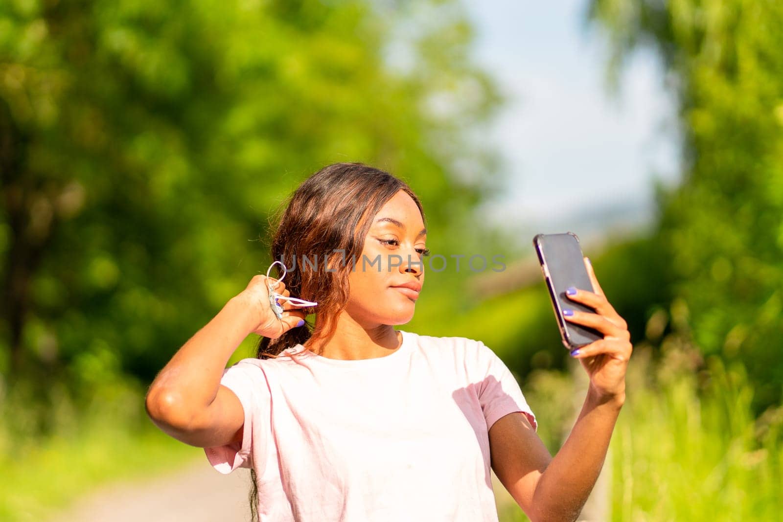 Young black woman with her phone taking selfie in a park on an October afternoon. by Ceballos