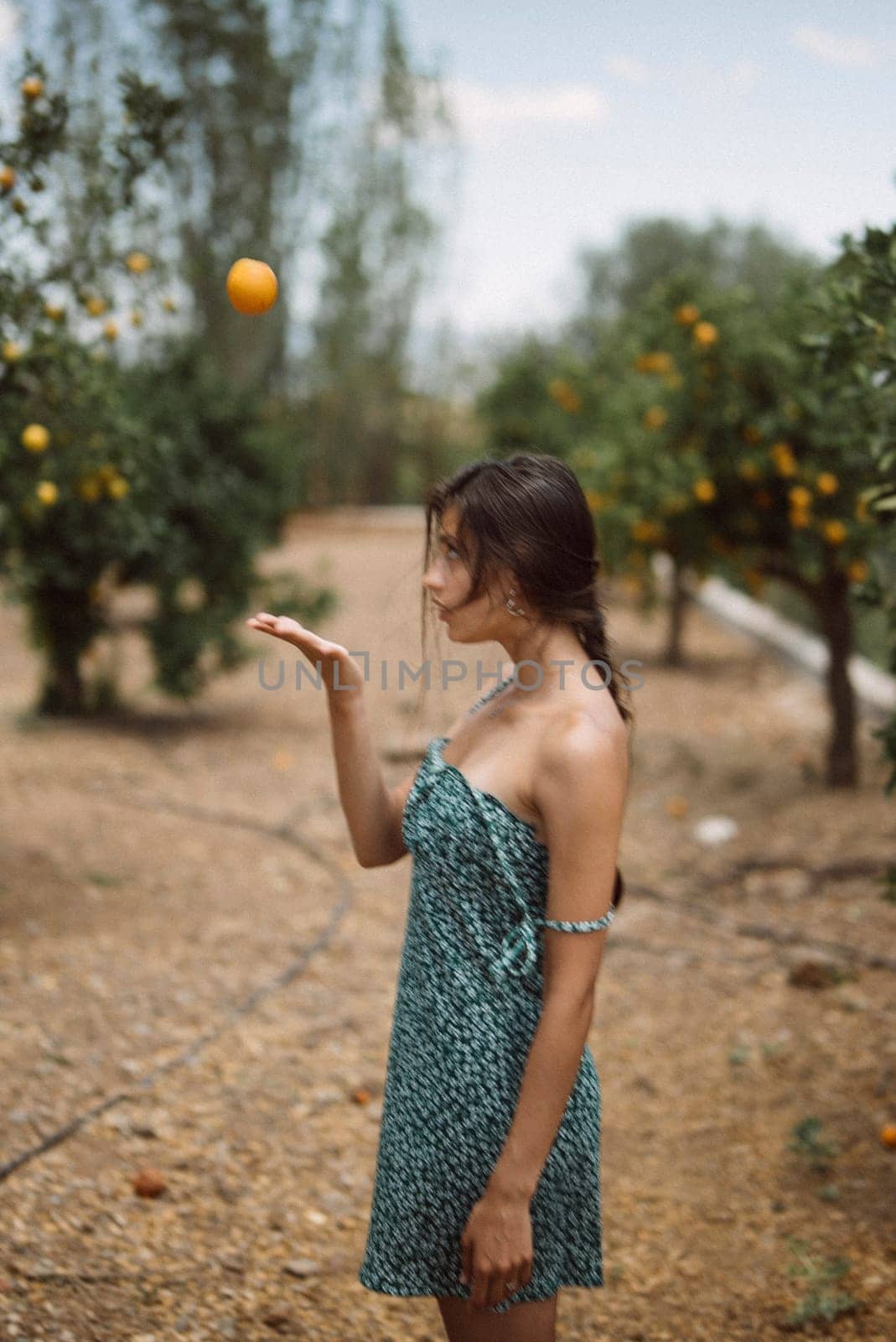A young woman wearing a green dress is playfully tossing an orange in the air while standing in a picturesque orchard