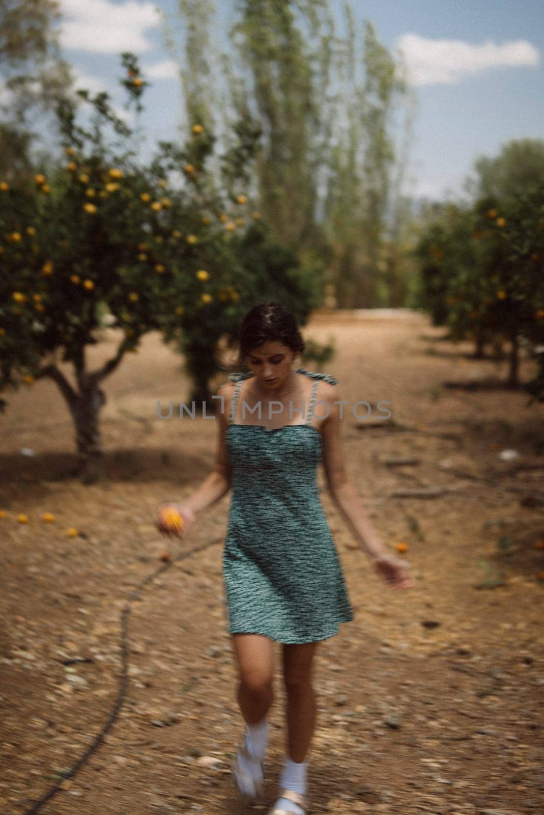 A young woman walks through an orchard on a sunny day, wearing a green dress and holding an orange fruit.